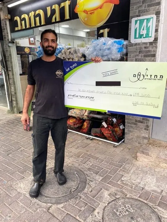 בעל עסק עומד מחוץ לחנות הדגים שלו ומחזיק צ'ק גדול על סך 17,250 ש"ח שקיבל כהחזר מס.